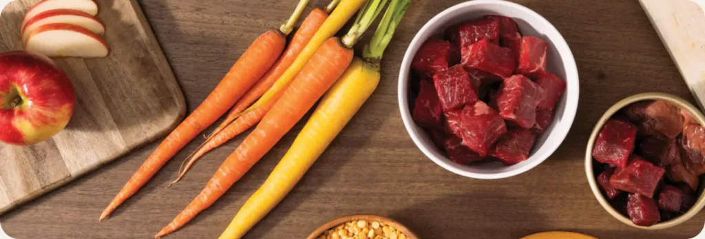 Carrots, apples and bowls of diced raw red meat on a wooden table